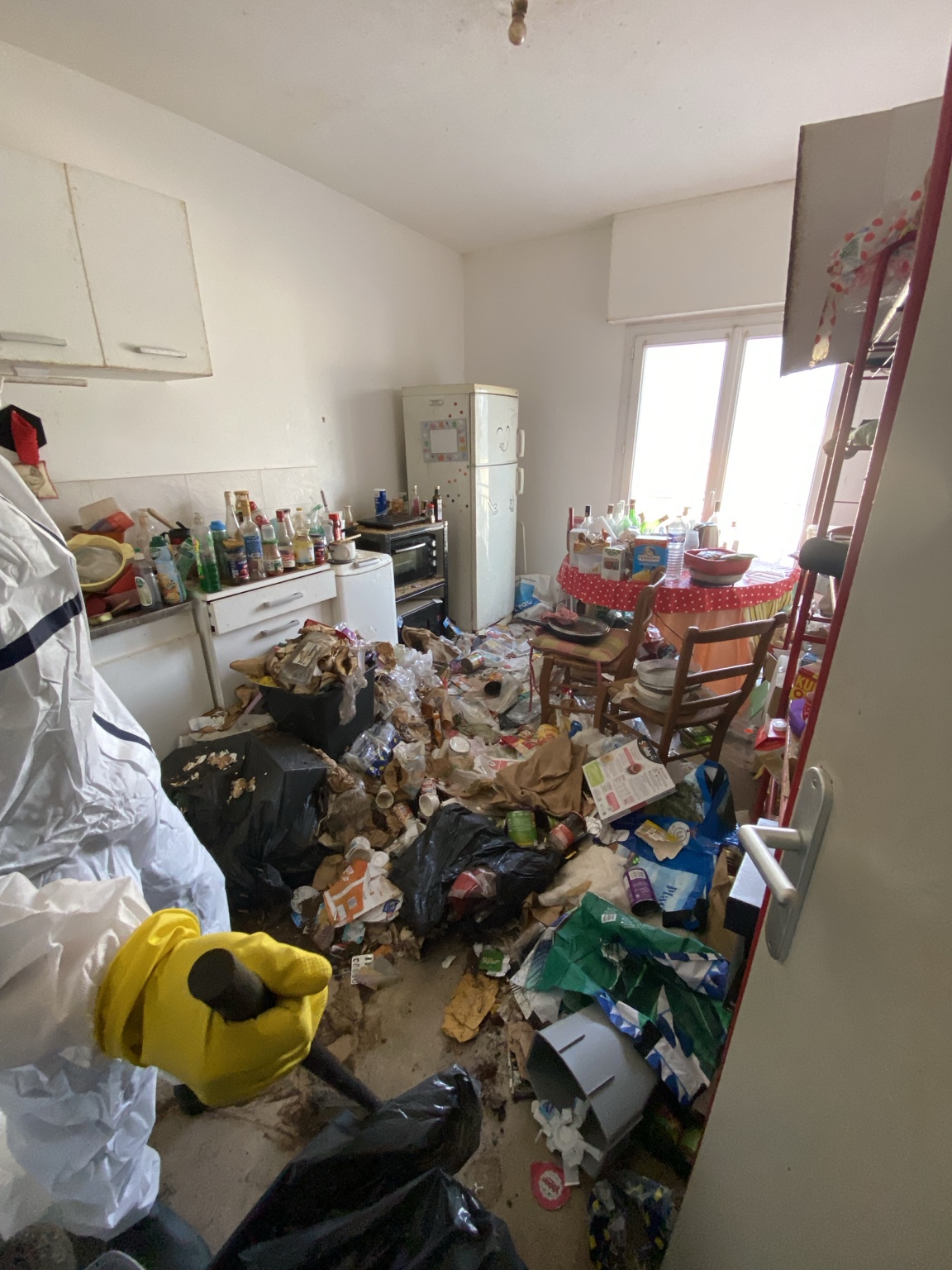 Vue d’une cuisine encombrée avec une personne en tenue de protection, visage hors cadre, au premier plan.